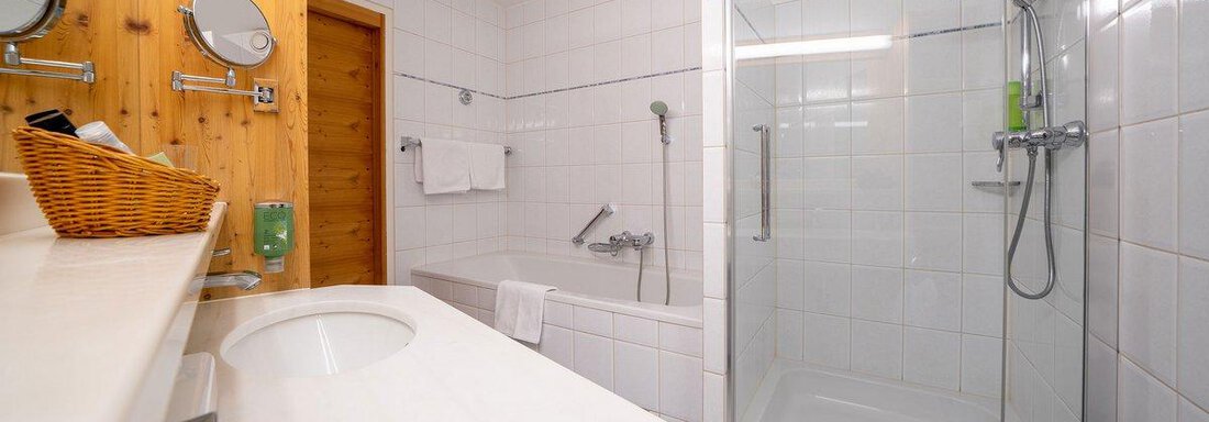 A modern bathroom with white tiles, a shower, and a bathtub. The wooden area gives the room a warm atmosphere. - Hotel Liebe Sonne - Sölden