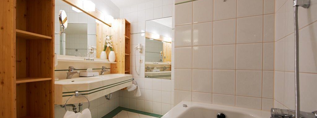 A modern bathroom with a bathtub and a large mirror. The walls are tiled in light colors, and there are wooden shelves for extra storage space. - Hotel Liebe Sonne - Sölden