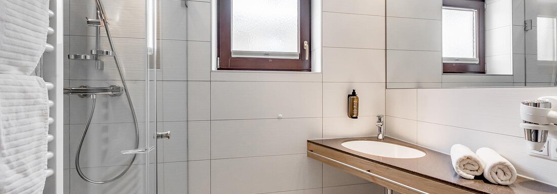 A modern bathroom with a glass shower enclosure and a bright tiled floor. Fresh towels are ready on the wooden shelf. - Hotel Sportpension Carinthia - Sölden