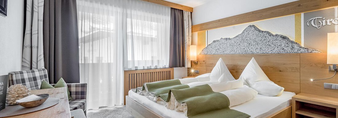 A modern bedroom with a large bed and stylish wooden decoration. Bright curtains and a cozy armchair can be seen in front of a window. - Hotel Sportpension Carinthia - Sölden