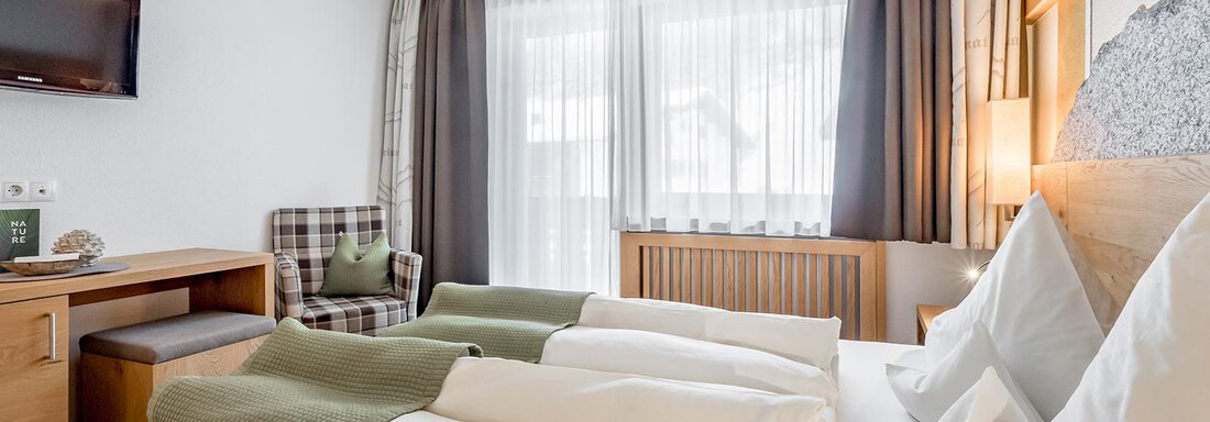 A modern hotel room with a double bed and fresh bedding. The room features a television, a cozy armchair, and large windows with plenty of natural light. - Hotel Sportpension Carinthia - Sölden