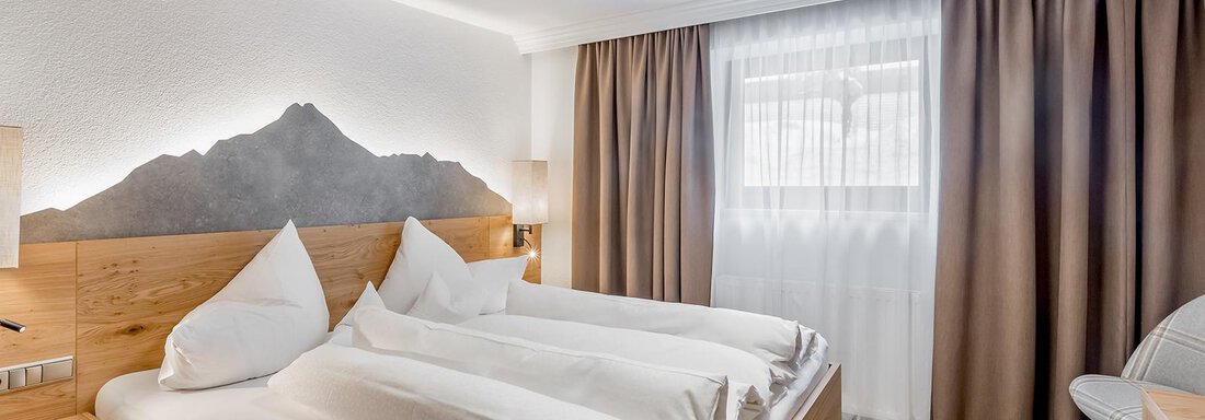 A modern hotel room with a large double bed and wooden tone furniture. The room is bright and friendly, with brown curtains and a beautiful view of the mountains. - Hotel Sportpension Carinthia - Sölden
