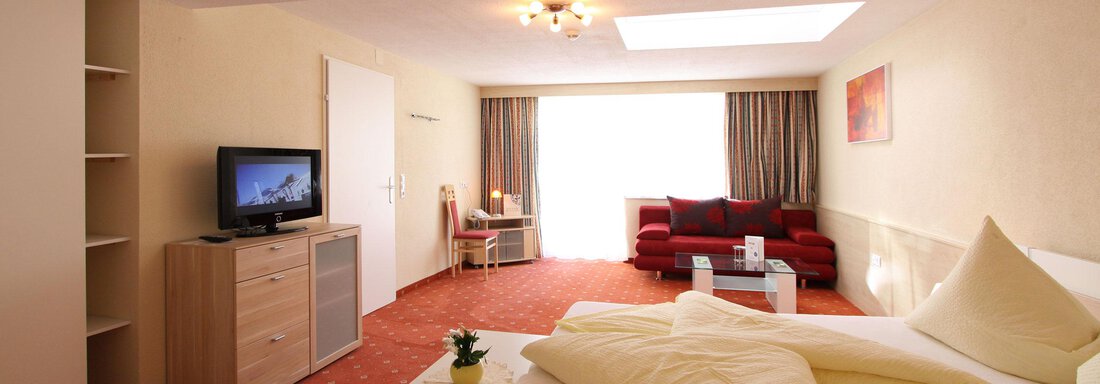 A modern room with a double bed, a TV, and a cozy red sofa. Large windows provide plenty of daylight. - Hotelappart Peter - Sölden