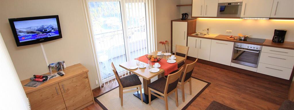 A modern kitchen with a dining area, featuring a table and chairs. In the background, there is a television on the wall and large windows that let in plenty of daylight. - Hotelappart Peter - Sölden