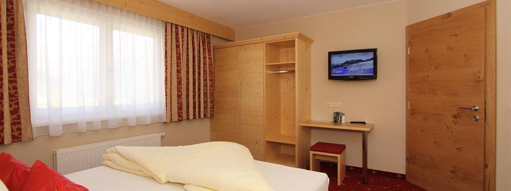 A cozy hotel room with a double bed, a television, and a workspace. The walls are light, and there are large windows that let in daylight. - Hotelappart Peter - Sölden