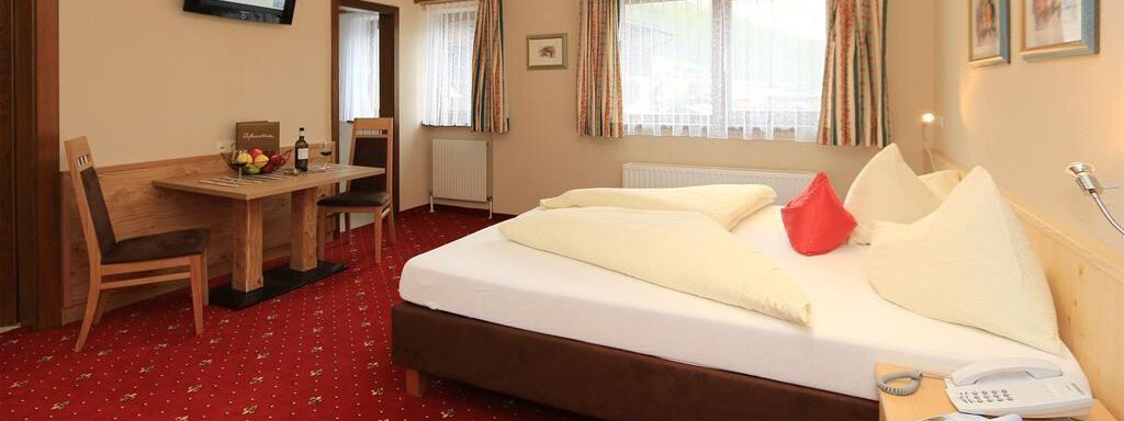 A cozy room with a large bed and red carpet. There is a small table with chairs and a television on the wall. - Hotelappart Peter - Sölden