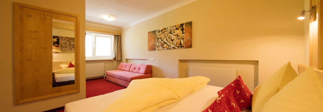 A cozy hotel room with a double bed and a separate sleeping area. The walls are painted in warm colors, and there is a large mirror as well as a sofa. - Hotelappart Peter - Sölden