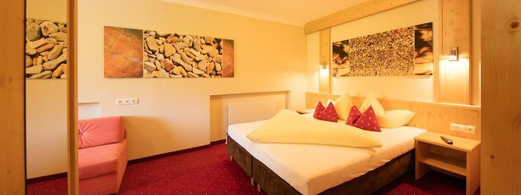 A cozy hotel room with a large bed and red cushions. The walls are decorated with pictures and there is a sitting area with a pink sofa. - Hotelappart Peter - Sölden