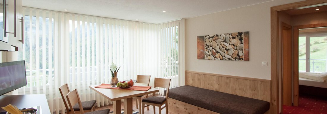 A bright living room with a dining table, chairs, and a sofa corner. Large windows provide plenty of daylight and offer a view of the outside. - Hotelappart Peter - Sölden