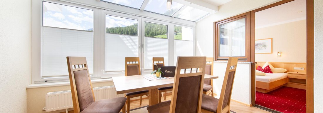 Wohnzimmer TYP 5 A bright dining area with a large table and wooden chairs. Light pours in through large windows, offering a view outside. - Hotelappart Peter - Sölden