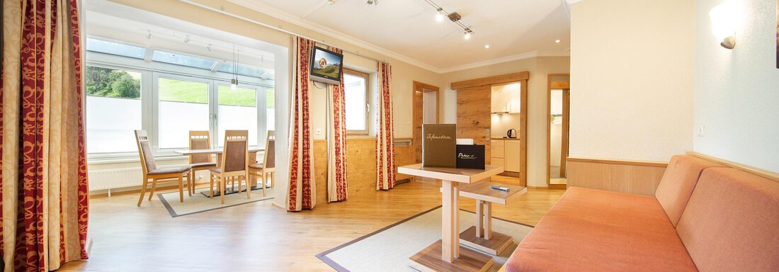 A bright, modern apartment with wooden flooring and large windows. The living area has a comfortable sofa and a dining area with tables and chairs. - Hotelappart Peter - Sölden