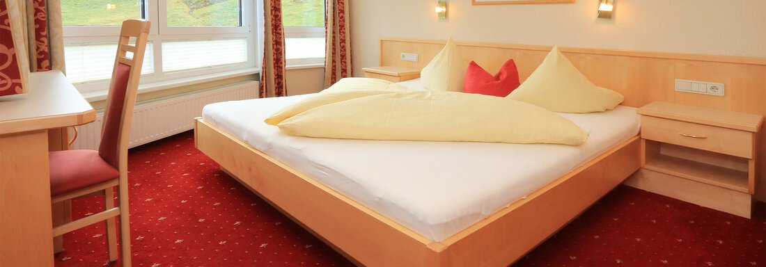 A cozy bedroom with a large bed and light wooden furniture. The walls are decorated with red curtains and the room has plenty of daylight. - Hotelappart Peter - Sölden