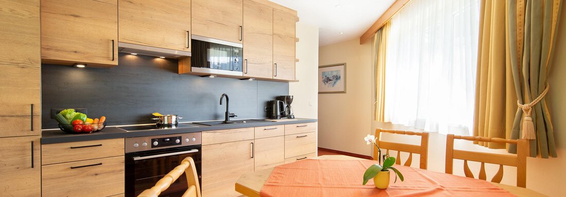 Wohnküche A modern kitchen with wooden cabinets and an energetic design. On the table, there is an orange tablecloth and a flower stands in the center. - Hotelappart Peter - Sölden