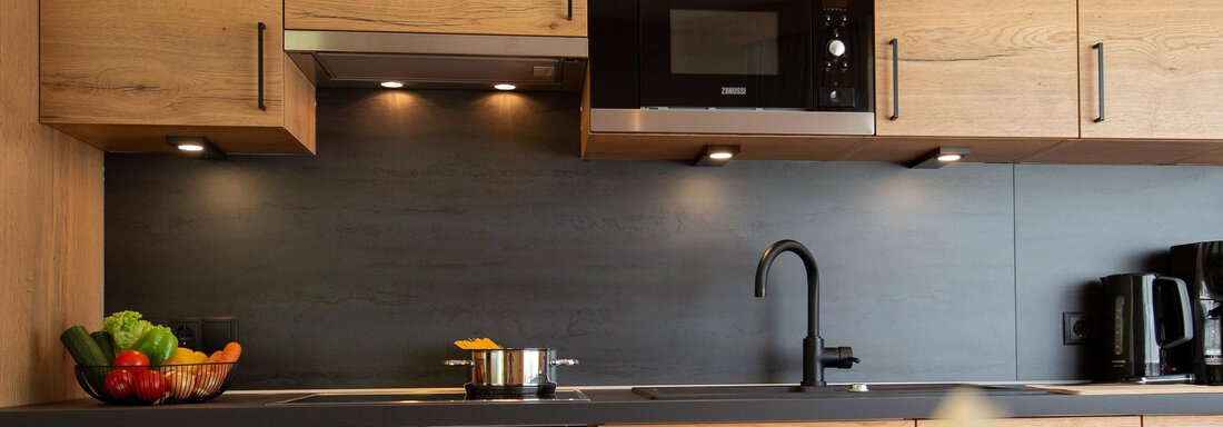 A modern kitchen with wooden furniture and a black countertop. On the work surface, there is fruit and a cooking area. - Hotelappart Peter - Sölden