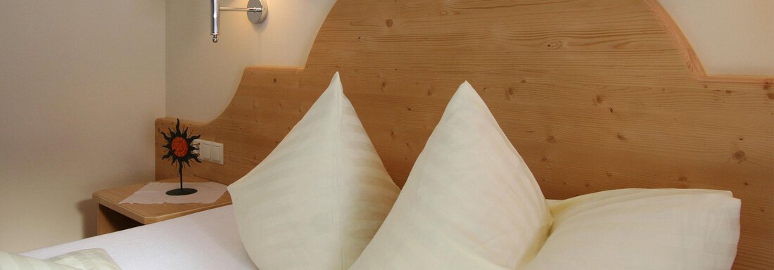 A cozy bed with white bedding and large, pillow-shaped pillows. On the wall, there is a wooden panel and a small lamp. - Hotelappart Peter - Sölden