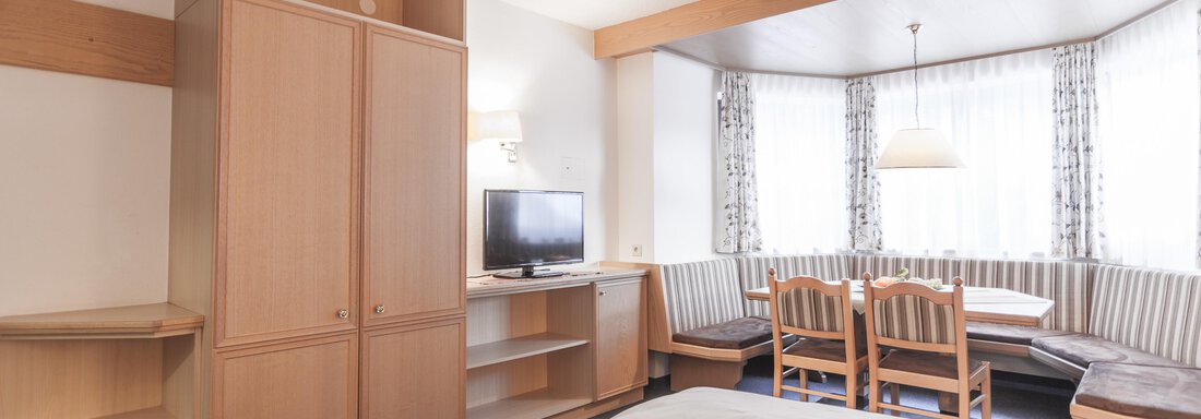 A bright hotel room with wooden furniture and a window seat. There is a television and a dining table with chairs. - Landhaus Falkner - Sölden