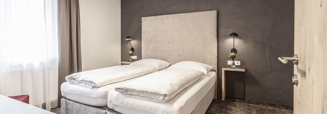 A modern bedroom with two separate beds and a stylish headboard. Next to the bed is a nightstand with a lamp, and the room features large windows with daylight. - Landhaus Falkner - Sölden