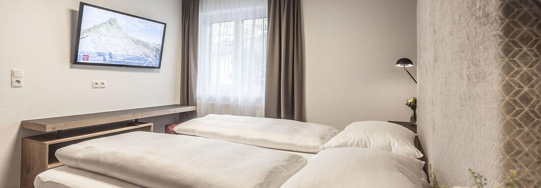 A modern hotel room with two single beds and a television on the wall. Large windows allow plenty of daylight in, and the room is tastefully furnished. - Landhaus Falkner - Sölden