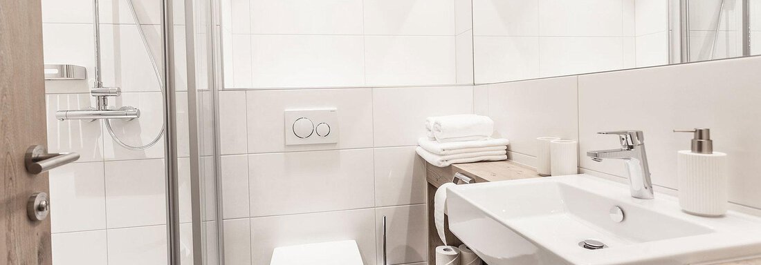 A modern bathroom with white tiles and clean lines. It includes a sink, a toilet, and a shower. - Landhaus Falkner - Sölden