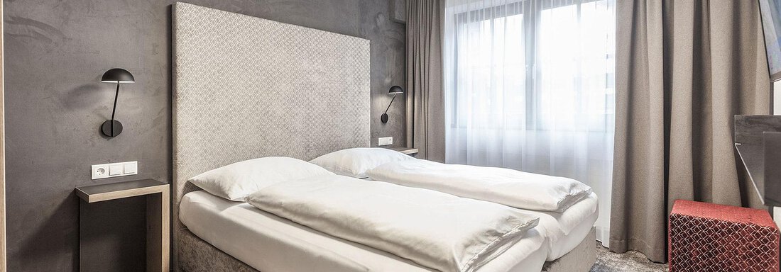 A modern bedroom with two single beds and a large window. The walls are in neutral colors, complemented by stylish curtains and simple lighting. - Landhaus Falkner - Sölden