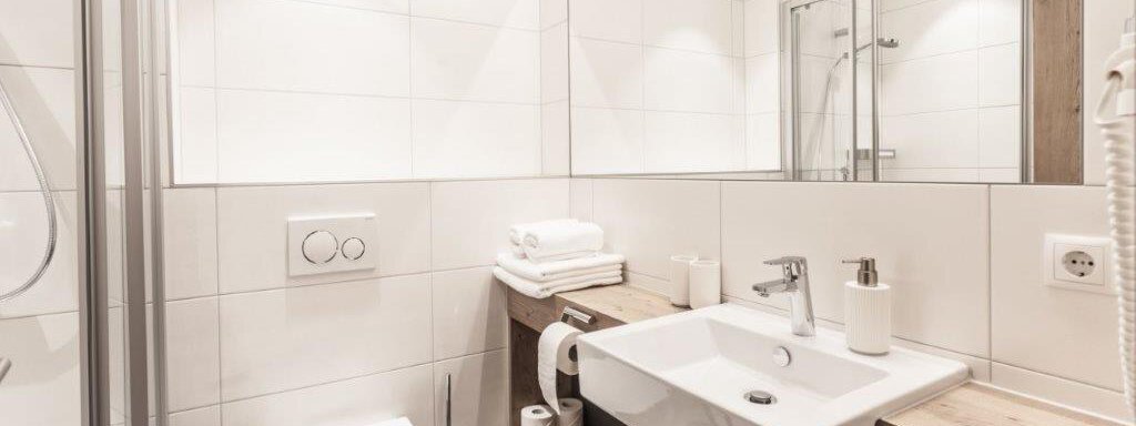 A modern bathroom with white tiles and a large mirror. It features a shower, a sink, and a toilet. - Landhaus Falkner - Sölden