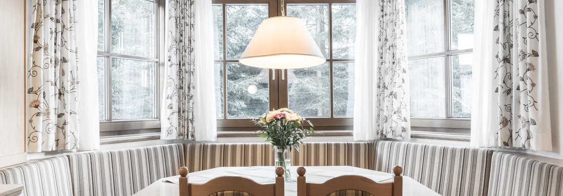 A cozy dining area with a round table and benches. Large windows with curtains let in plenty of light. - Landhaus Falkner - Sölden