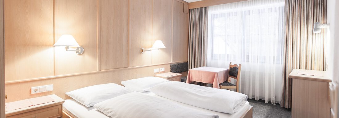 A cozy hotel room with two beds and wooden paneling. The room features large windows with curtains and a small table. - Landhaus Falkner - Sölden