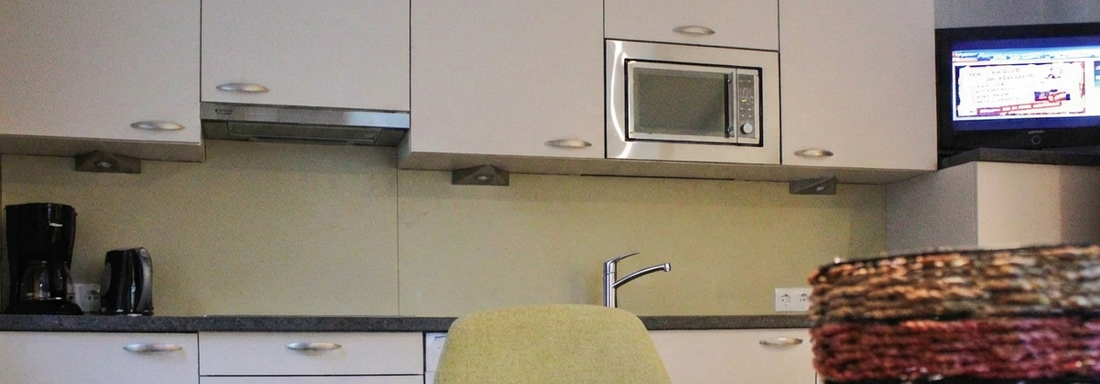 A modern kitchen with bright cabinets and a microwave. On the table, there is a place mat, while a television is visible in the background. - Lutonia Appartements Sölden - Sölden