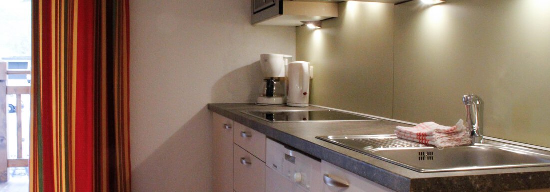 A modern, well-equipped kitchen with bright cabinets and a sink. Red and yellow curtains create a cozy atmosphere. - Lutonia Appartements Sölden - Sölden