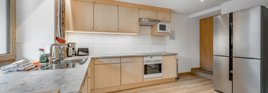 A modern kitchen with bright wooden cabinets and an elegant granite countertop. There is a refrigerator and a microwave as well as large windows that provide plenty of light. - Lyns Home - Sölden