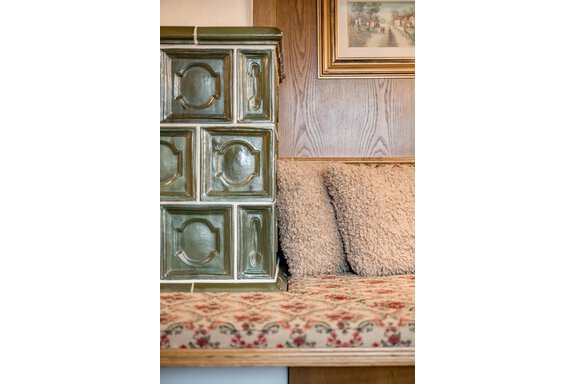 A rustic room with a decorative green stove and a cozy bench. The bench is upholstered with a floral fabric and soft cushions. - Lyns Home - Sölden