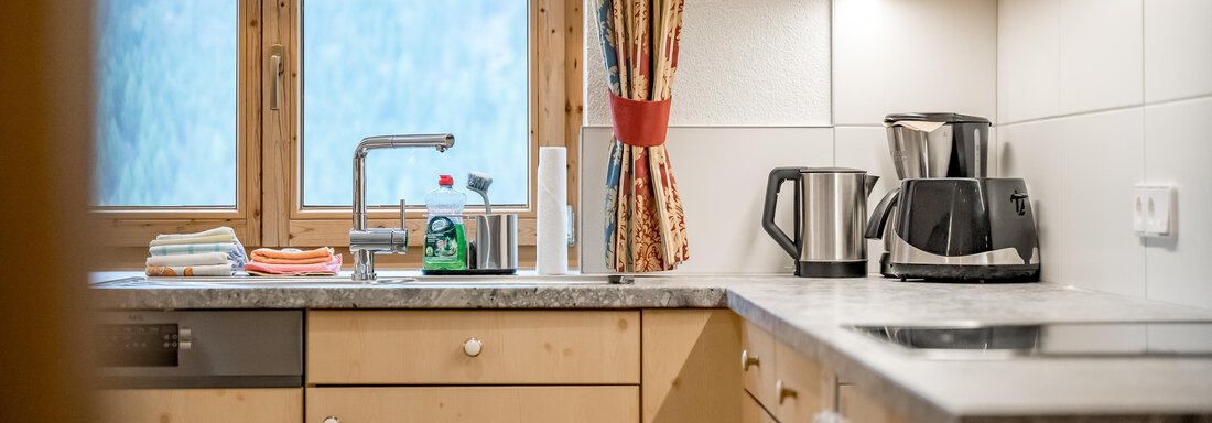 A modern, bright kitchen with wooden cabinets and a window that lets in daylight. Various kitchen appliances and utensils are placed on the countertop. - Lyns Home - Sölden
