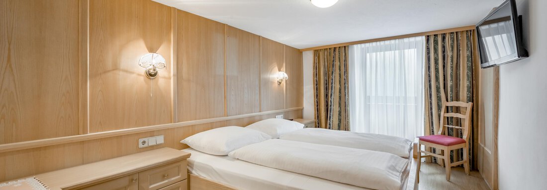 A modern bedroom with two single beds and cozy wooden paneling. There is a window with curtains and a television on the wall. - Lyns Home - Sölden
