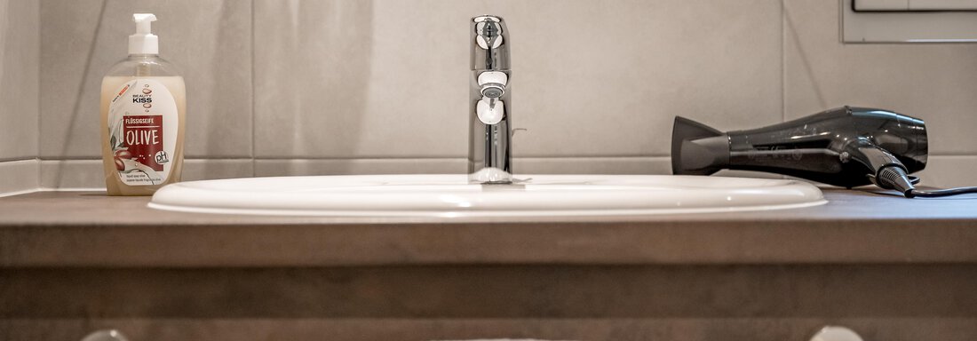 A modern bathroom with a sink and a hairdryer. Next to the sink is a bottle of hand soap and a towel is hanging on the towel rack. - Lyns Home - Sölden