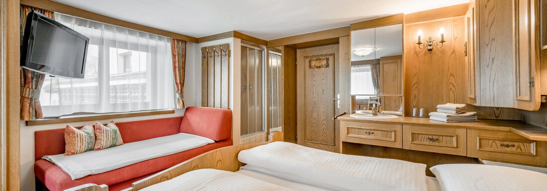 A cozy hotel room with two twin beds, a red sofa, and a television. The room features warm wood and beige tones, as well as large windows that ensure plenty of daylight. - Lyns Home - Sölden
