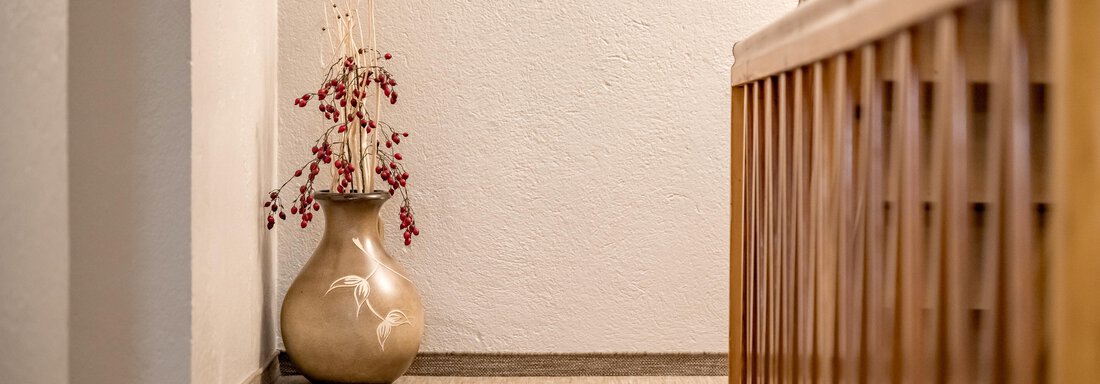 A hallway with a wooden railing and a carpet. In the corner stands a decorative vase with dried flowers. - Lyns Home - Sölden