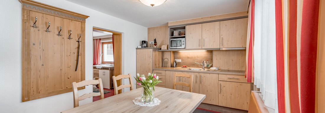 260221_130115 A cozy kitchen with a wooden table and chairs. The walls are light and the windows have red curtains. - Lyns Home - Sölden