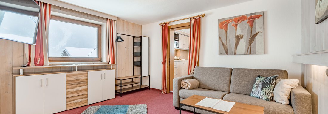 A cozy living room with a sofa, a table, and a colorful rug. The walls are bright, and large windows let in plenty of light. - Lyns Home - Sölden
