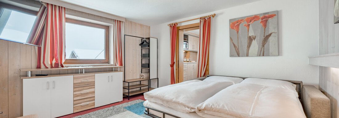 A cozy bedroom with a large double bed and a carpet. The walls are adorned with red curtains and a piece of artwork. - Lyns Home - Sölden