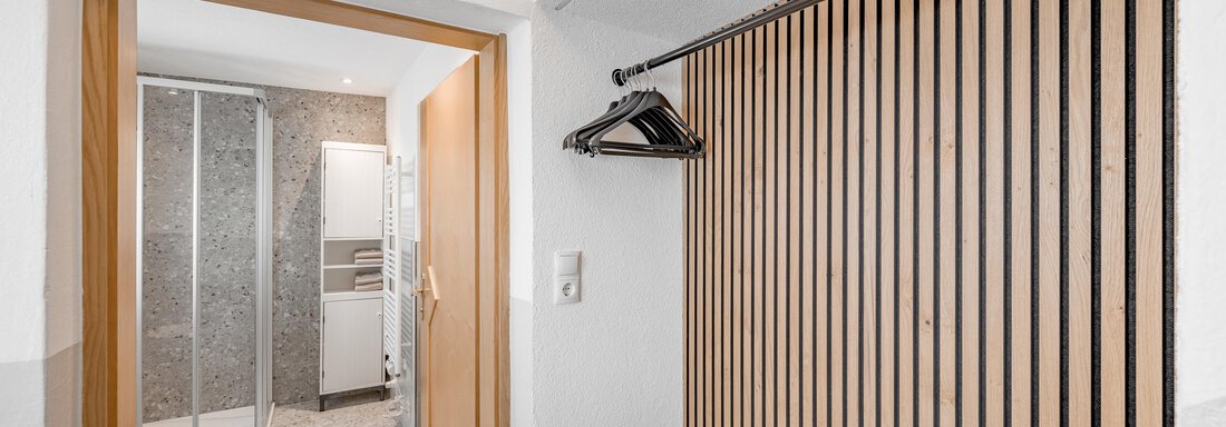 A modern hallway with a wooden coat rack and an open connection to a bathroom. The floor is light, and the space feels friendly and inviting. - Lyns Home - Sölden