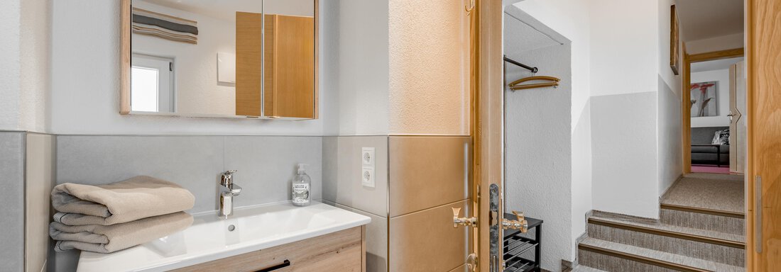 A modern bathroom with a wooden vanity and large mirrors. In the background, stairs and a bright hallway can be seen. - Lyns Home - Sölden