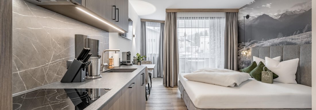 251126_160828 A modern kitchen with wooden cabinets and a cooking area. In the background, there is a cozy bedroom with a large bed and windows that let in plenty of light. - Marco's Mountain Home - Sölden