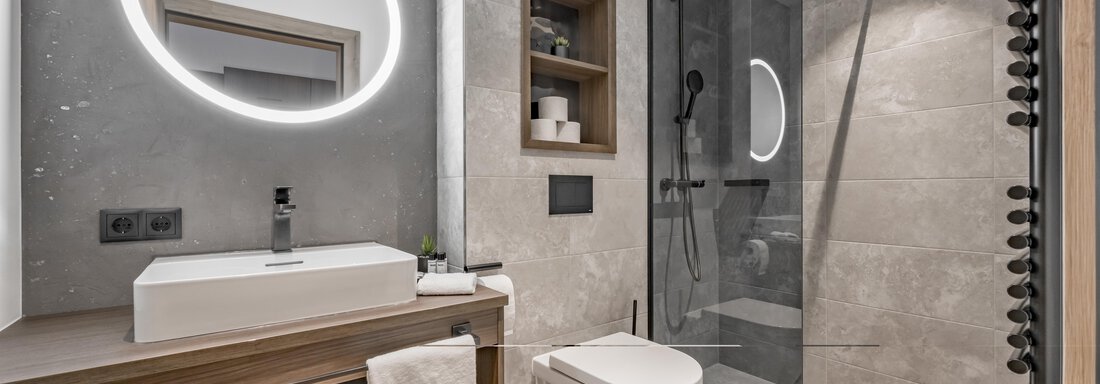 A modern bathroom with a glass shower cabin and a simple sink. The walls are painted in neutral gray tones, and a round mirror with lighting complements the design. - Marco's Mountain Home - Sölden