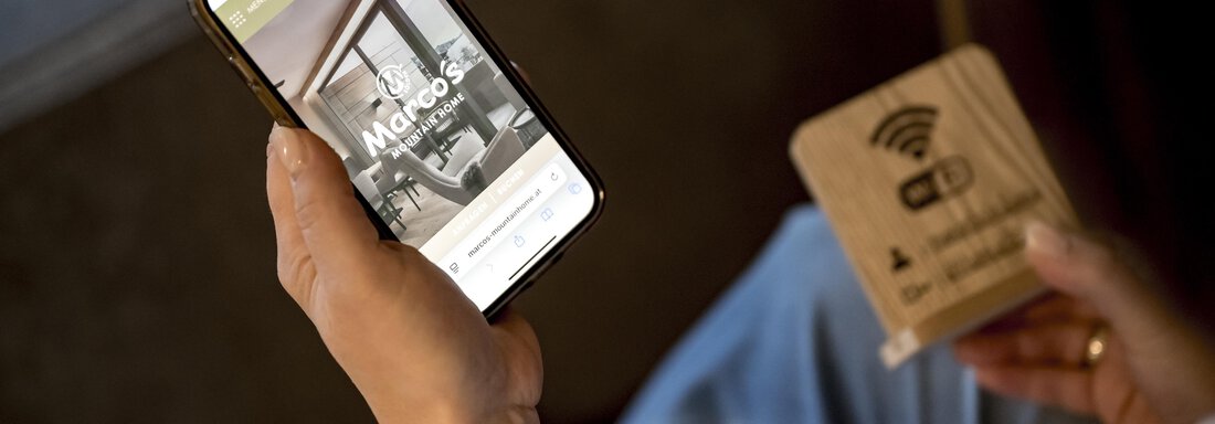 A person is holding a smartphone in their hand, displaying an app. Next to the phone is a wooden sign with a Wi-Fi symbol. - Marco's Mountain Home - Sölden