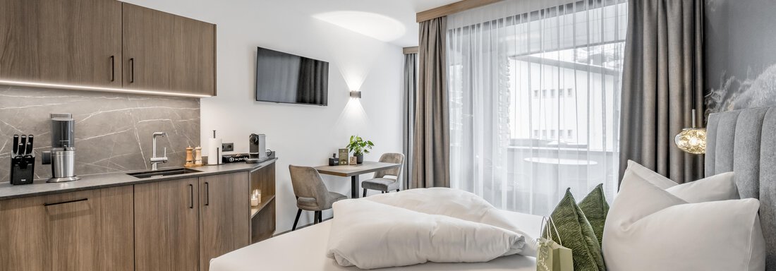 A modern room with a large bed, a small table, and a kitchenette.  
Large windows provide plenty of daylight. - Marco's Mountain Home - Sölden