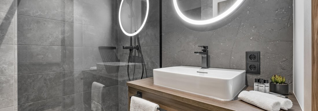 A modern bathroom with gray tiles and an elegant shower. The sink area is equipped with a wood-colored base cabinet and oval mirrors. - Marco's Mountain Home - Sölden