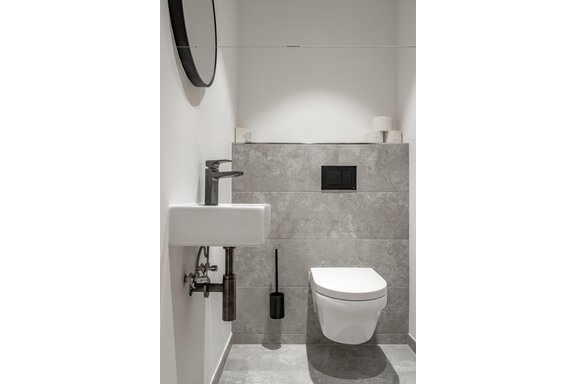 A modern bathroom with gray tiles and a white toilet. On the wall, there is a sink and a round mirror. - Marco's Mountain Home - Sölden