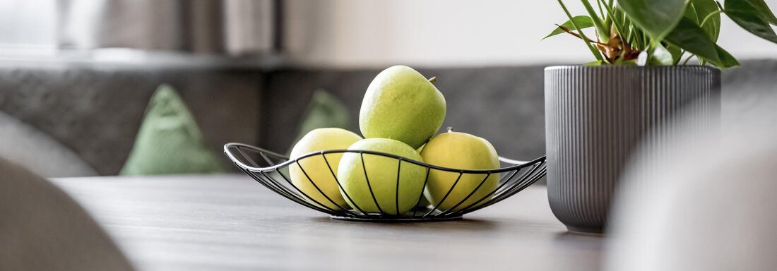 A dining table with a wire basket full of green apples and a pot with a plant. The background shows a cozy living atmosphere. - Marco's Mountain Home - Sölden
