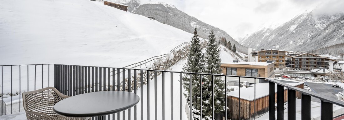 A balcony with a table and two chairs, from which one can view a snow-covered landscape. In the background, mountains and some buildings are visible. - Marco's Mountain Home - Sölden