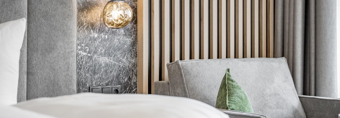 A modern hotel room with a gray sofa and a green decorative pillow. A warmly glowing lamp hangs over the bed. - Marco's Mountain Home - Sölden